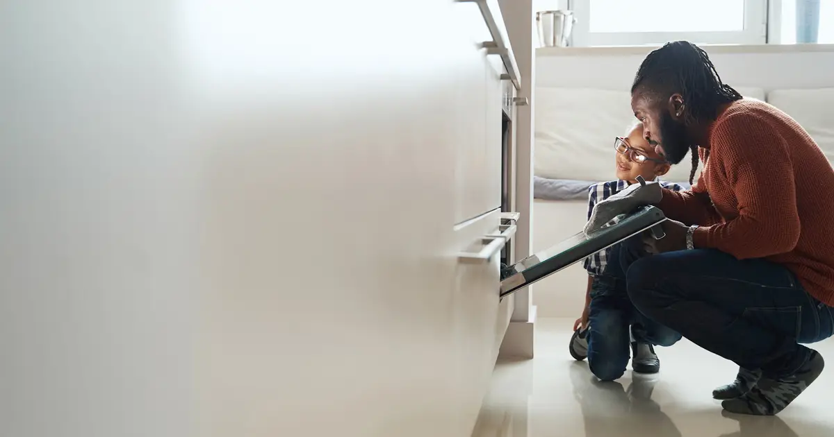 A man and a child are kneeling in front of an open oven. The man is wearing an orange sweater, gray pants, and an oven mitt, while the child, dressed in a plaid shirt and glasses, is smiling and looking at him. The scene appears warm and domestic, set in a modern kitchen.