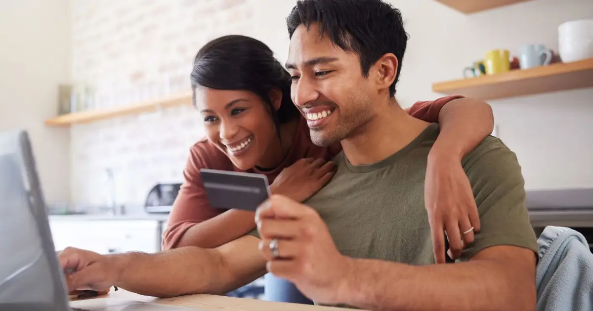 Smiling couple in kitchen using laptop and credit card together.