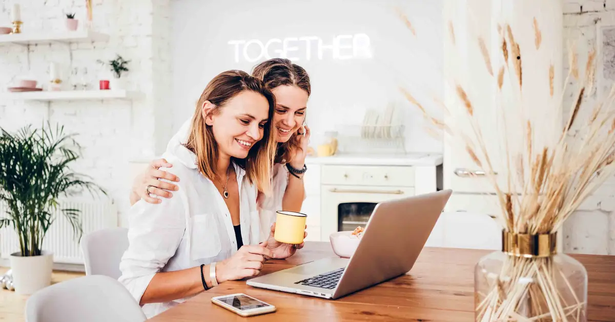 Two women smiling at laptop in cozy kitchen, one holding mug.