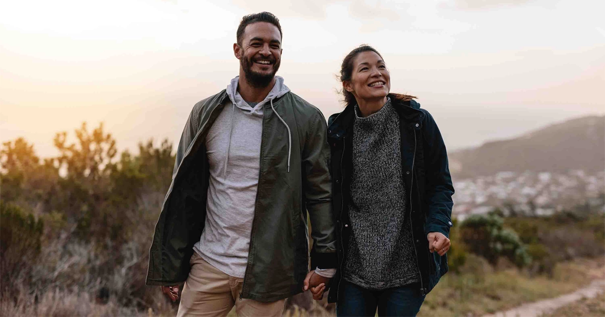 Smiling couple holds hands walking outdoors at sunset in casual jackets.