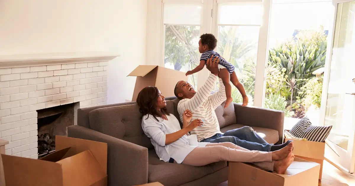 A joyful family relaxes in their new home, surrounded by moving boxes. The father lifts their baby into the air while the mother smiles beside him on the couch. Sunlight streams in through large windows, highlighting the bright and welcoming living space.